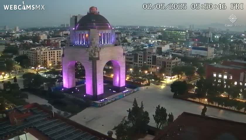 A large monument, illuminated in purple, stands in a city square at dusk, surrounded by city buildings and trees.
