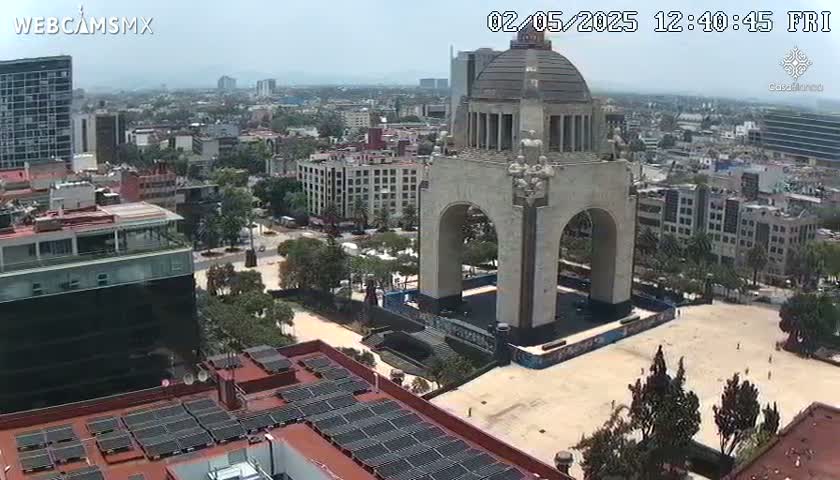 A high-angle view on a clear day shows a large monument with an archway in a city square surrounded by buildings, trees, and solar panels on rooftops.