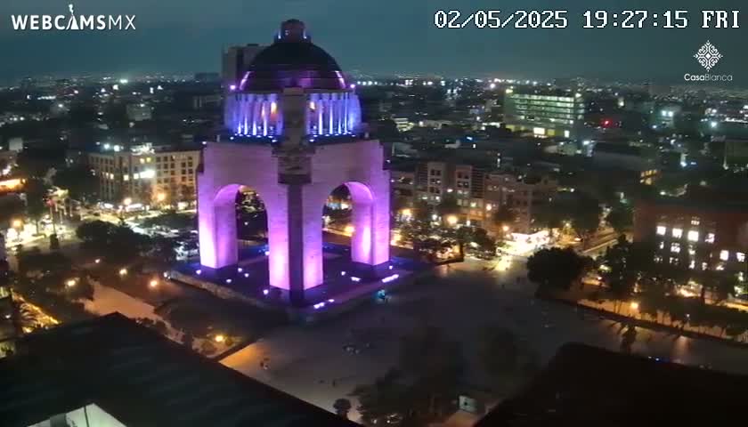 A large monument, illuminated in purple, is seen at night in a city with surrounding buildings and streets also lit up.
