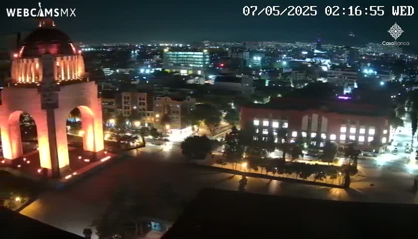 A nighttime, high-angle view shows a monument illuminated in orange, surrounded by a city with buildings and streets lit up.