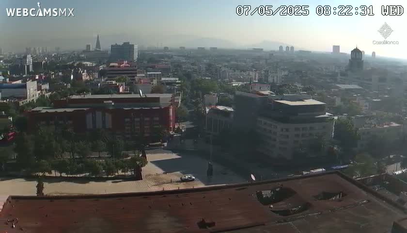 A hazy, sunny day view of a sprawling city from a high vantage point, showing numerous buildings and a large square.