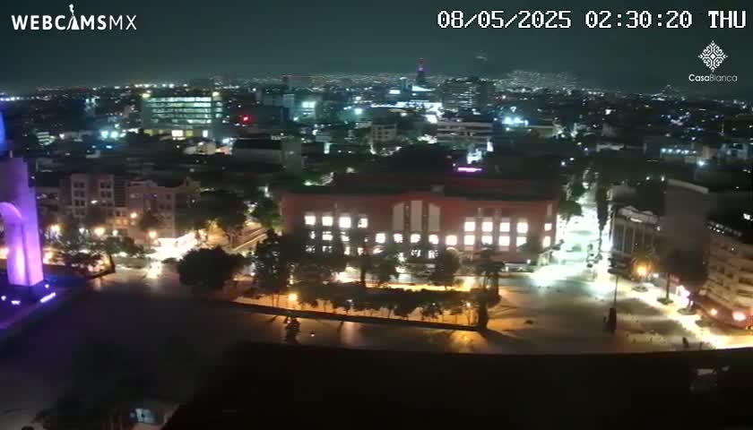 A nighttime aerial view of a city, showing brightly lit buildings and streets, with a large plaza in the foreground.