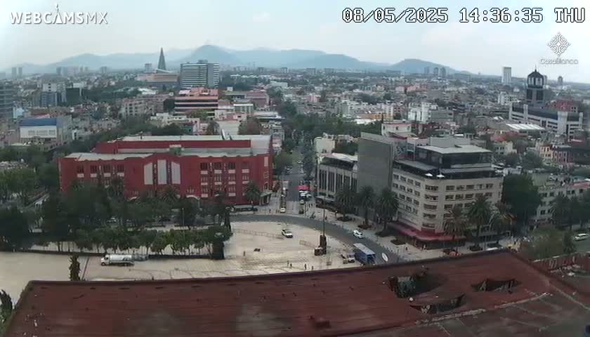 An overcast view of a city square and surrounding buildings, showing a mostly empty plaza with a few vehicles and some construction, under a hazy sky.
