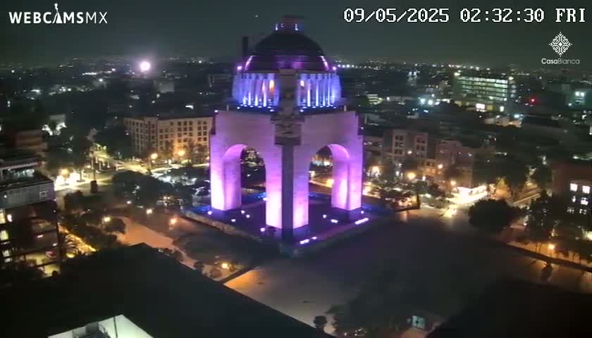 A large, illuminated monument stands in a city at night, surrounded by other buildings and streets.