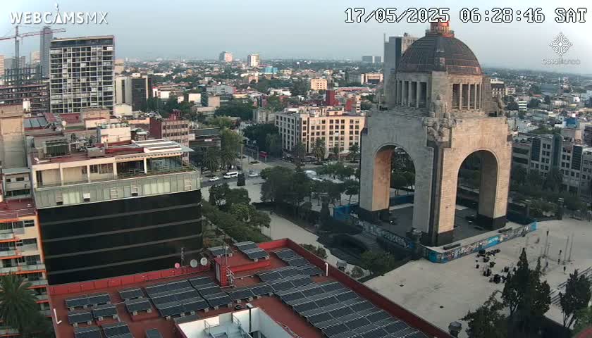 An aerial view of a city on a partly cloudy day, showing a large monument with an archway in a plaza surrounded by buildings and trees.