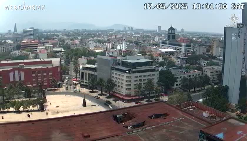 A sprawling city scene under a hazy sky shows numerous buildings of varying heights and styles, with palm trees lining some streets and a large plaza in the foreground.