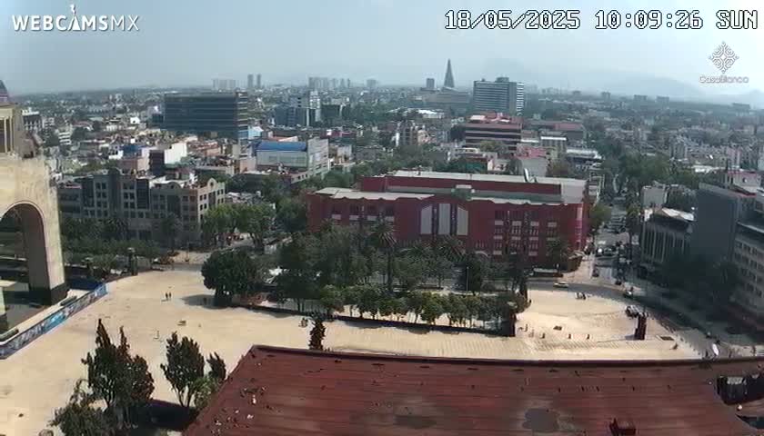 A high-angle, long shot on a sunny day shows a large city square with a reddish-brown building and surrounding cityscape, including high-rise buildings in the background.