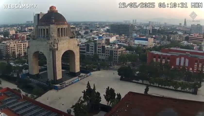 A large stone monument with an archway stands in a plaza, overlooking a city on a hazy, overcast day.