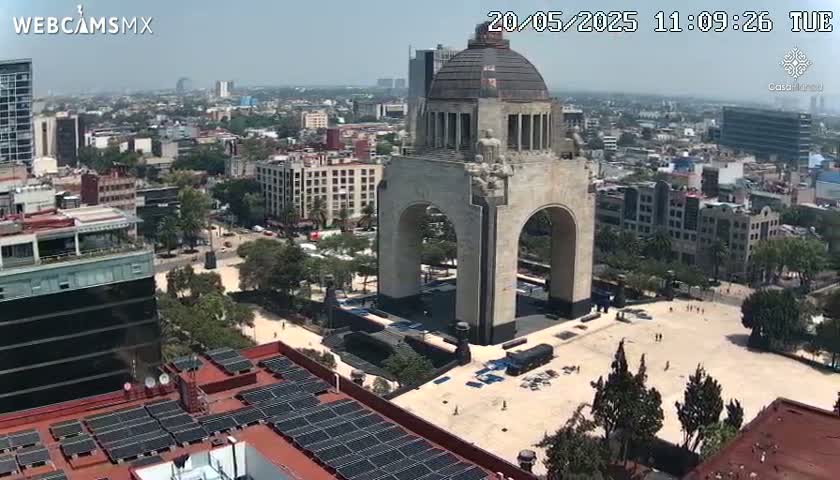 A large monument with an archway stands in a city square on a sunny day, surrounded by buildings and trees.