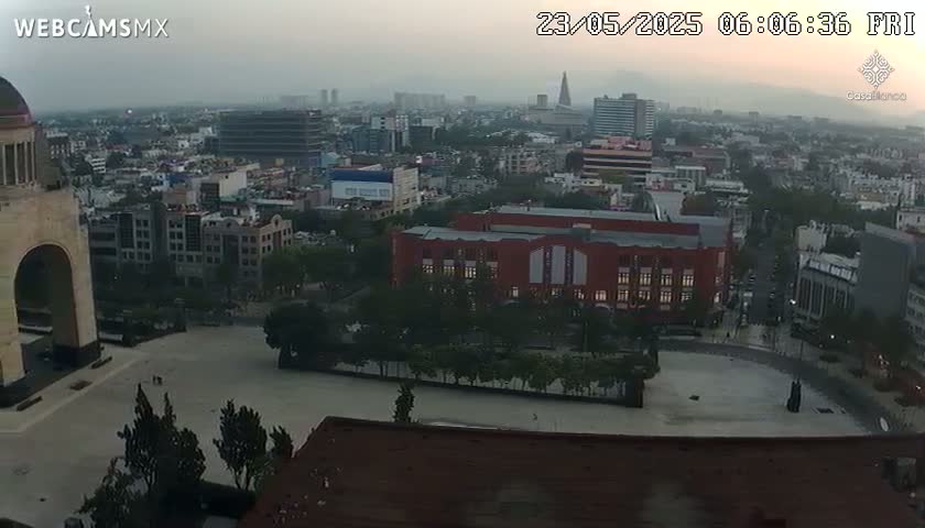 A hazy sunrise over a large city reveals a wide plaza in front of a monumental arch, with a large brick building visible in the mid-ground.