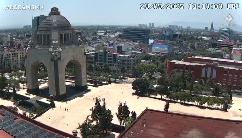 A large stone monument with arches stands in a plaza surrounded by a city under a clear, sunny sky.