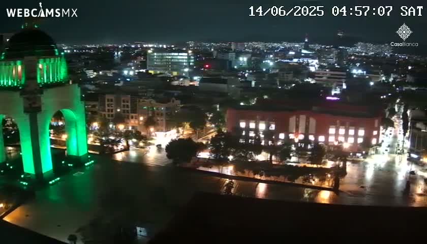 A nighttime aerial view of a city, showing a large monument illuminated in green light, and surrounding buildings and streets.