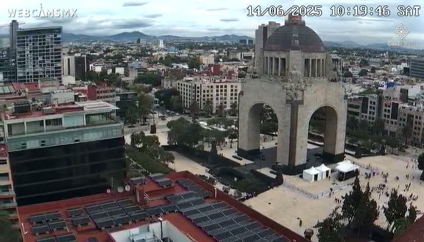 An aerial view on an overcast day shows a large monument with an archway in a city square, surrounded by buildings and trees.