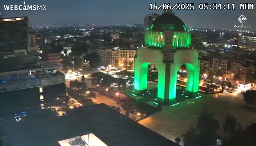 A nighttime aerial view of a city shows a large monument illuminated in green light, surrounded by buildings and streets with minimal traffic.