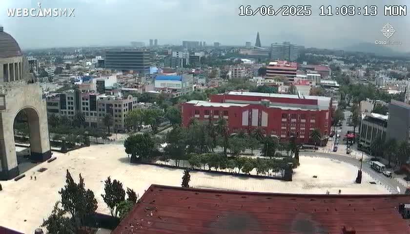 A high-angle view of a city square on a partly cloudy day, showing a large plaza surrounded by buildings, trees, and a portion of a monumental arch.