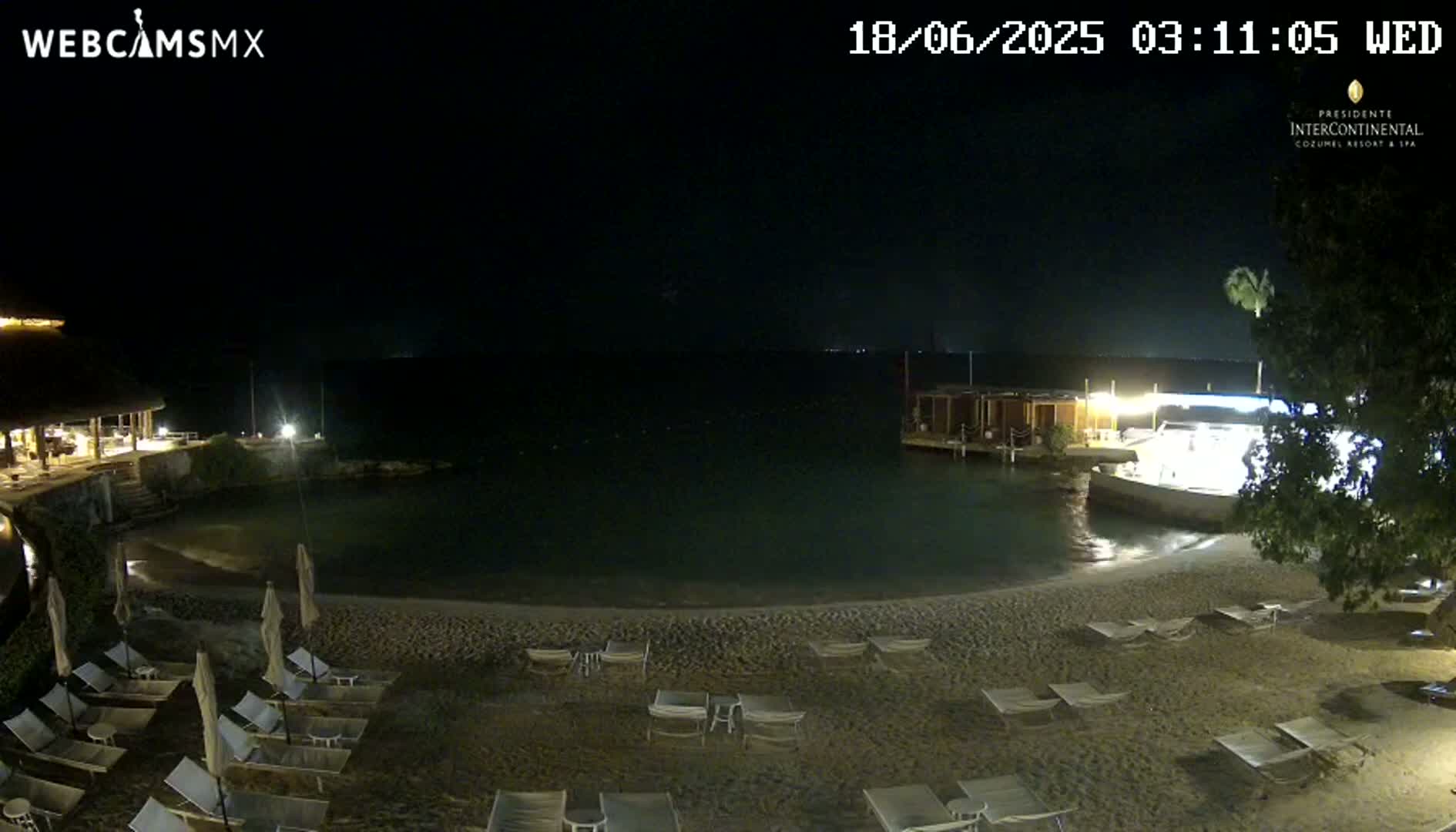 A dark, nighttime beach scene shows empty lounge chairs on the sand, a calm body of water, and a building in the background.
