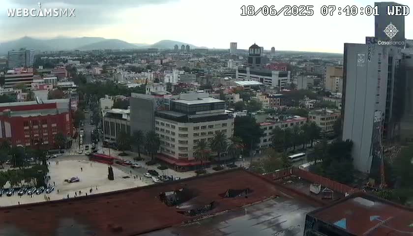 A high-angle view of a city under overcast skies shows a large plaza, numerous buildings, and distant mountains.