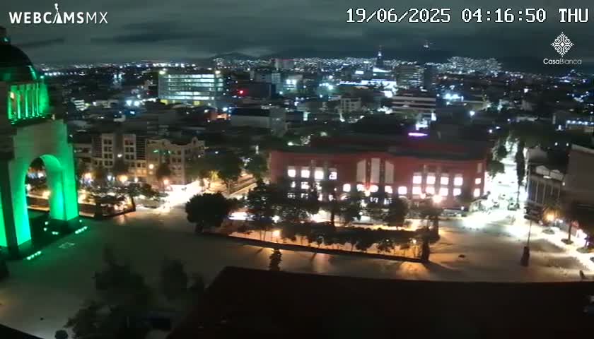 A nighttime aerial view of a city illuminated by lights, showing a large monument glowing green, and a large building in the foreground.