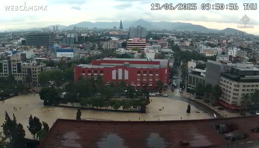 A high-angle, wide shot of a city on an overcast day shows a large plaza partially flooded with water, surrounded by buildings, trees, and a large red building in the center.
