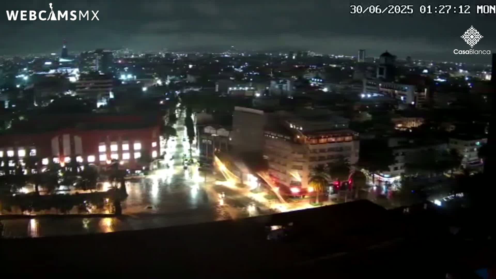 A nighttime aerial view of a city experiencing heavy rain, with flooded streets and illuminated buildings.