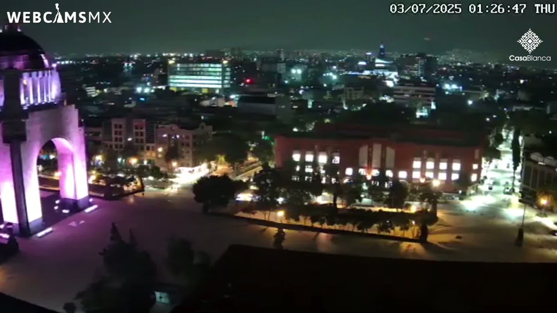 A nighttime, high-angle view shows a brightly lit monument in a city square, surrounded by buildings and trees under a clear sky.
