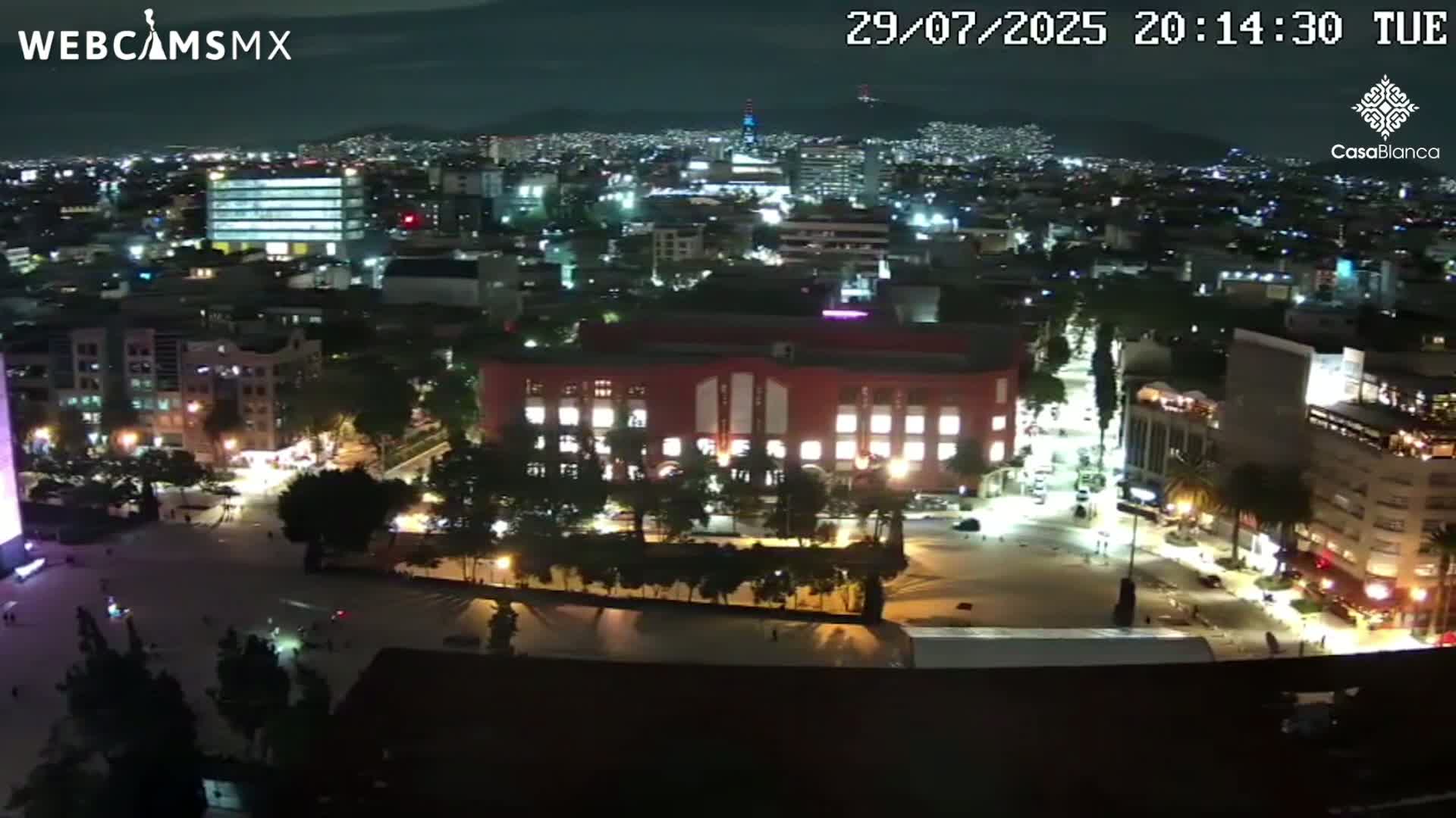 A nighttime aerial view of a city with brightly lit streets and buildings under a hazy sky.