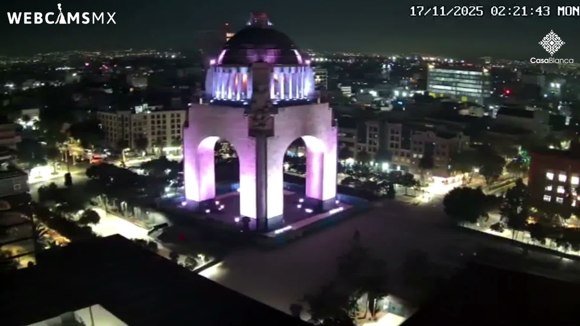 A nighttime aerial view reveals a grand monument glowing with purple lights, surrounded by a sprawling city of illuminated buildings and streets under a clear sky.