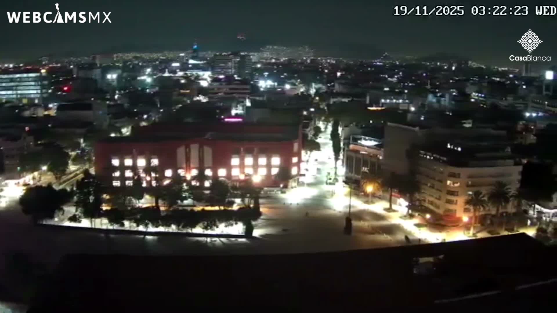 A clear night reveals a city skyline from an elevated perspective, featuring a brightly lit red-orange building in the midground and an expansive urban landscape twinkling with lights under a dark sky.