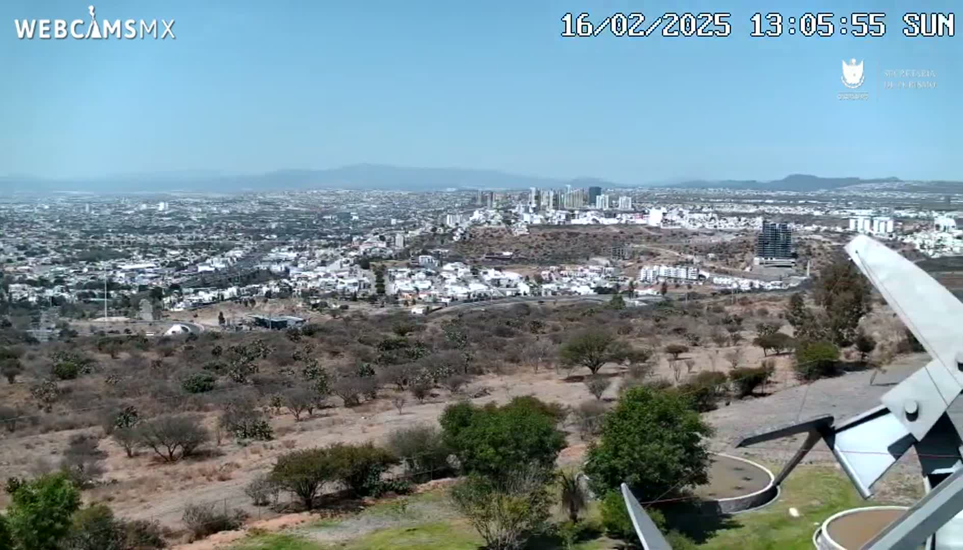 A sprawling city under a clear, sunny sky is viewed from a high vantage point, with scrubby vegetation and a portion of a metal structure in the foreground.