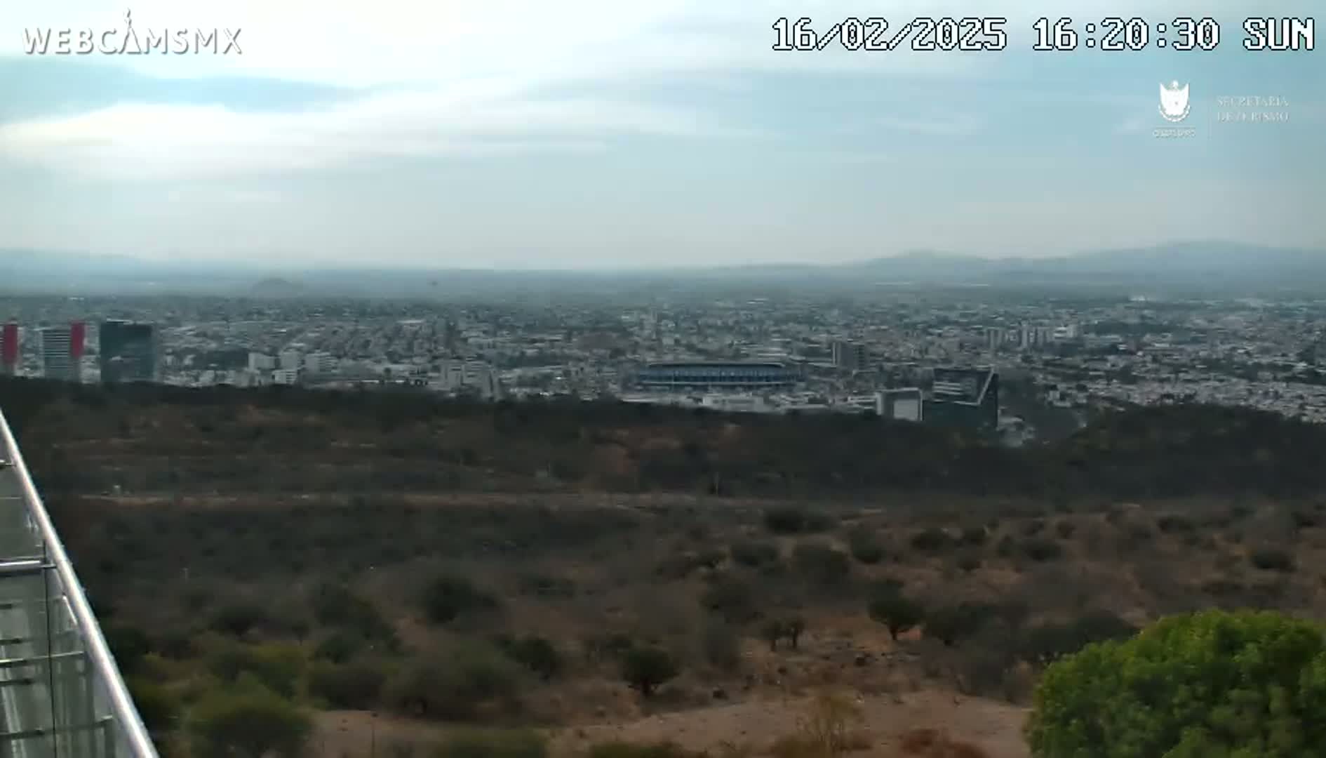 A hazy view from a hilltop shows a sprawling city with a stadium and tall buildings in the distance, under a mostly cloudy sky.