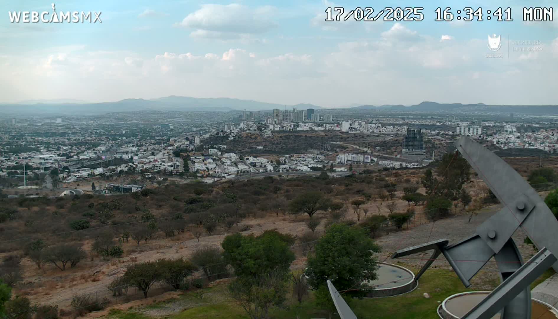 A partly cloudy day reveals a sprawling city view from a high vantage point, featuring a large metal sculpture in the foreground.