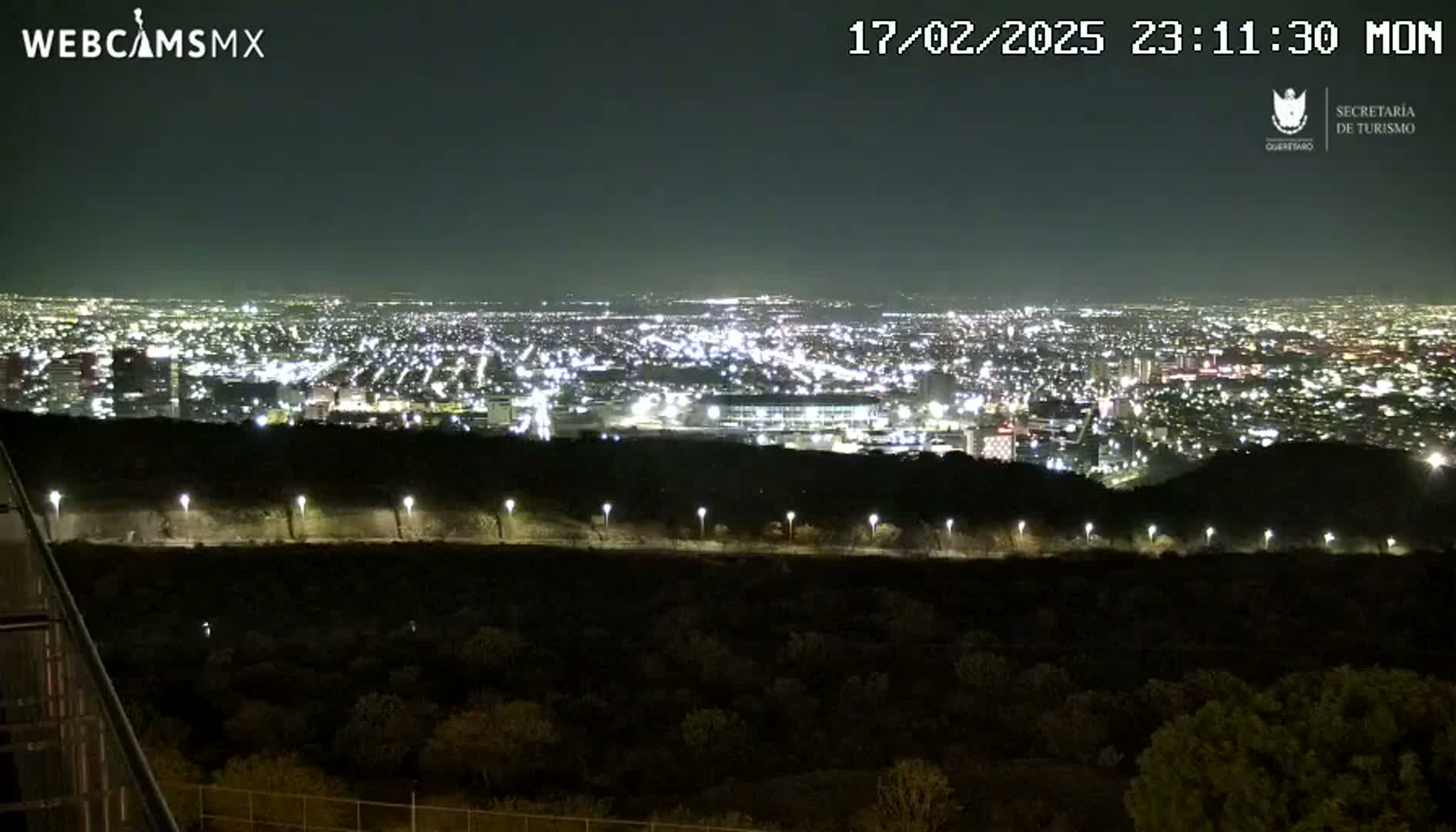 A nighttime view from an elevated position shows a sprawling city illuminated by countless lights, with a dark hillside and sparsely lit road in the foreground under a clear sky.