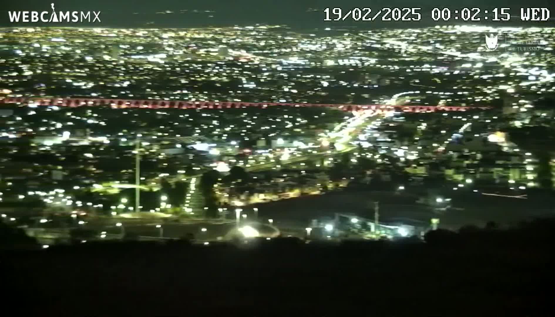A nighttime, high-angle view shows a sprawling city illuminated by countless lights, with roads and highways visible below.
