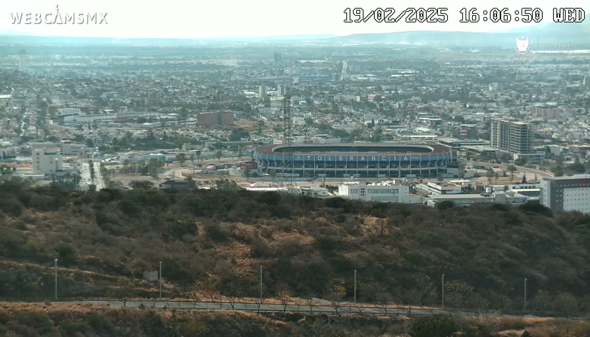 A long shot from a hilltop reveals a sprawling city under a hazy sky, featuring a stadium in the midground and dry, scrubby vegetation in the foreground.