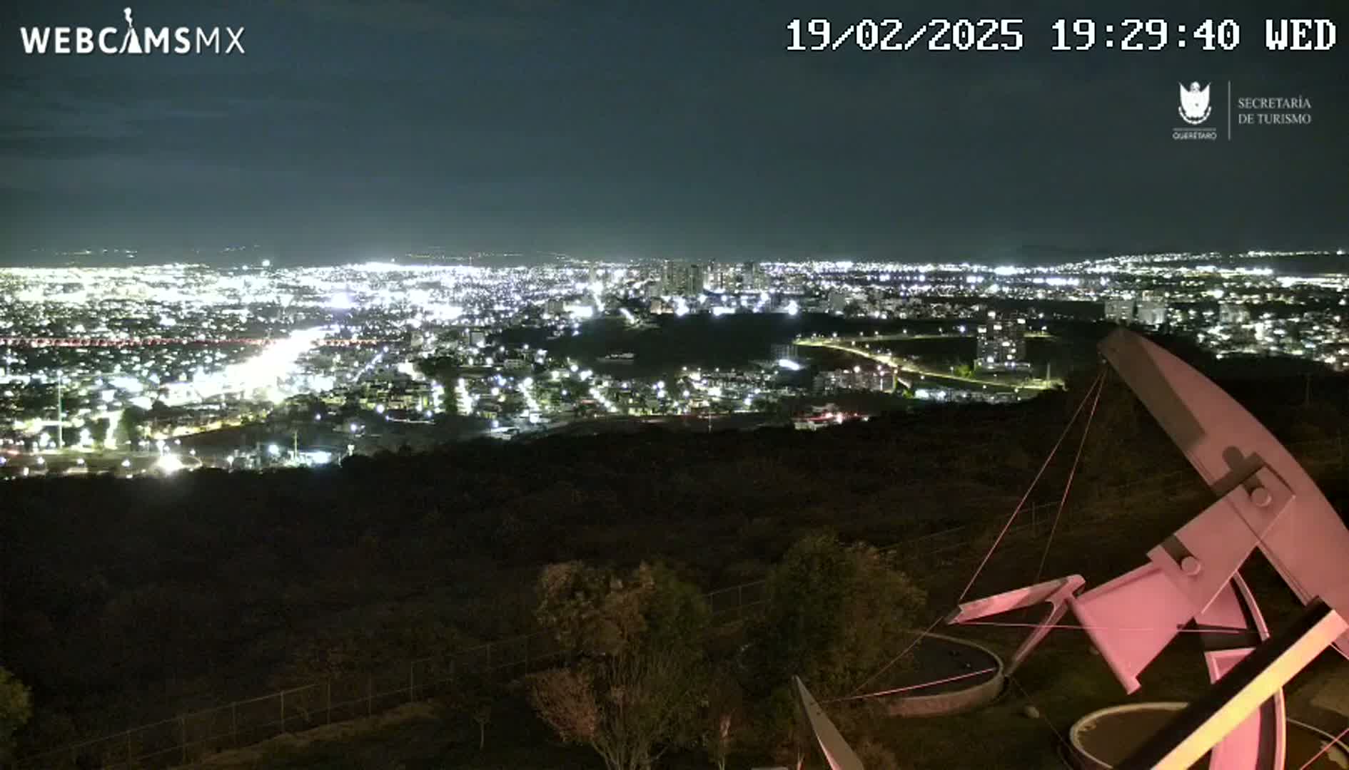 A nighttime view from an elevated vantage point shows a sprawling city illuminated below, with a large, pinkish-white metal structure in the foreground.