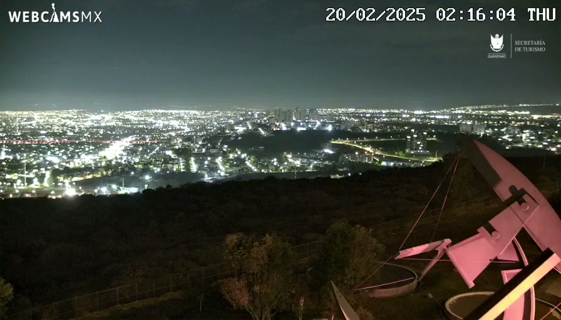 A nighttime view from an elevated vantage point shows a sprawling city illuminated below, with a large, angular pink structure in the foreground under a clear sky.