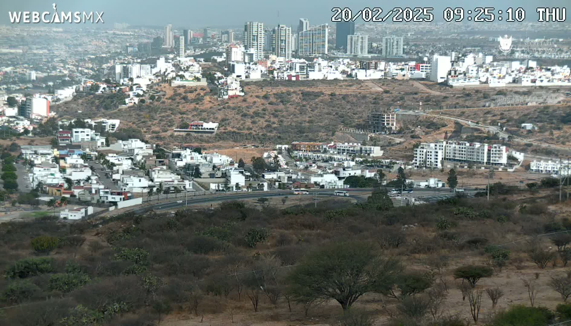 A hazy, high-angle view shows a sprawling city with numerous white buildings, a road curving through a scrubby, dry landscape below, and taller buildings in the distance.