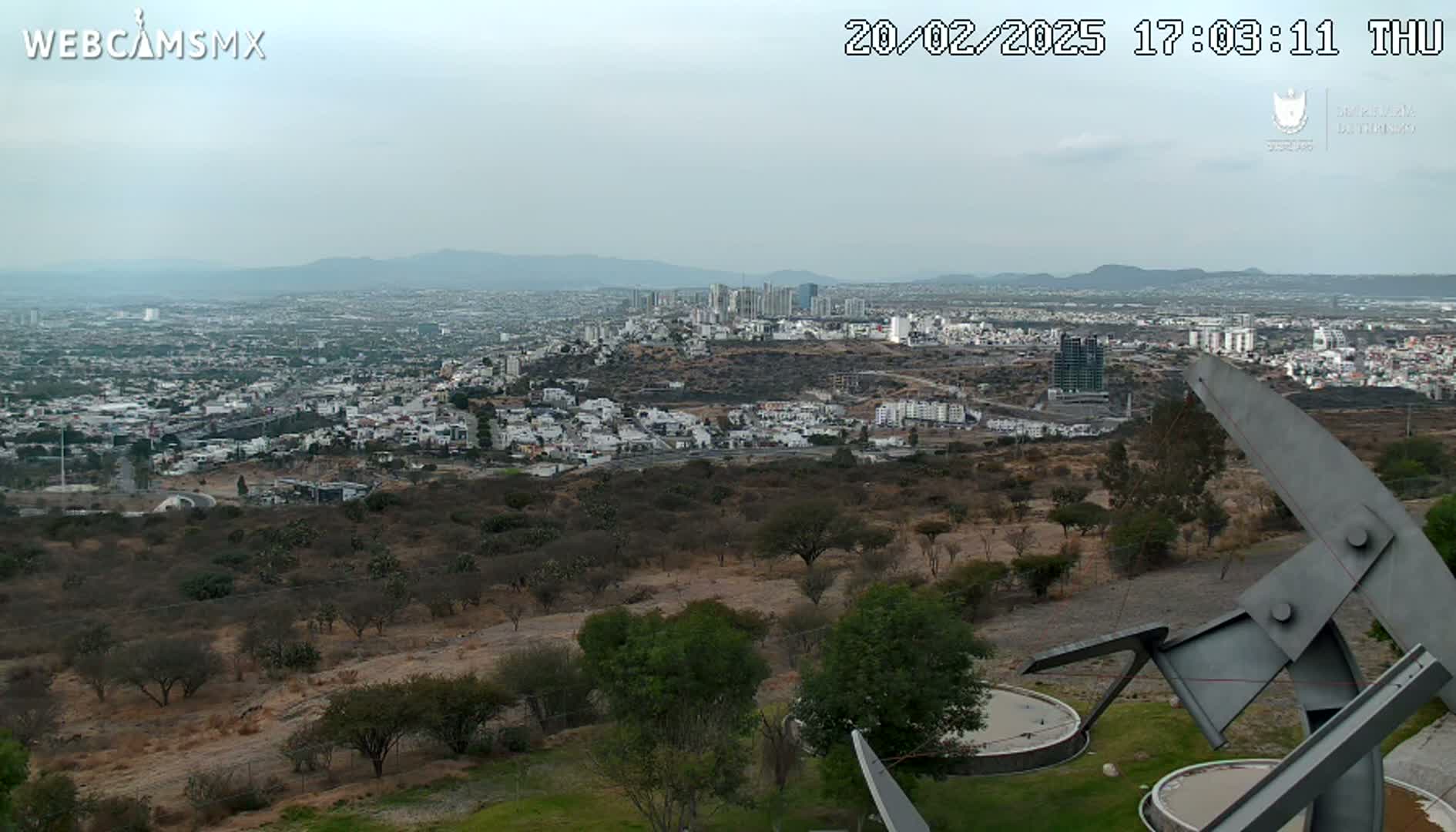 A sprawling city under a hazy sky is viewed from a hilltop, with a large metal sculpture in the foreground.