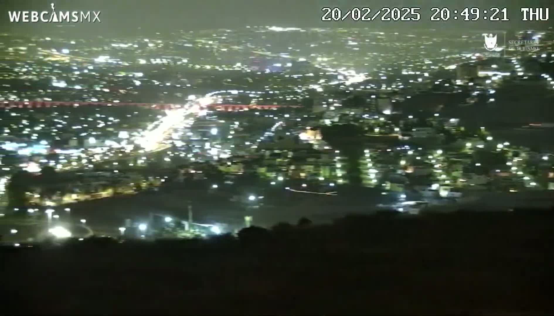 A nighttime, high-angle view shows a sprawling city illuminated by numerous lights, with a slightly hazy or cloudy atmosphere.