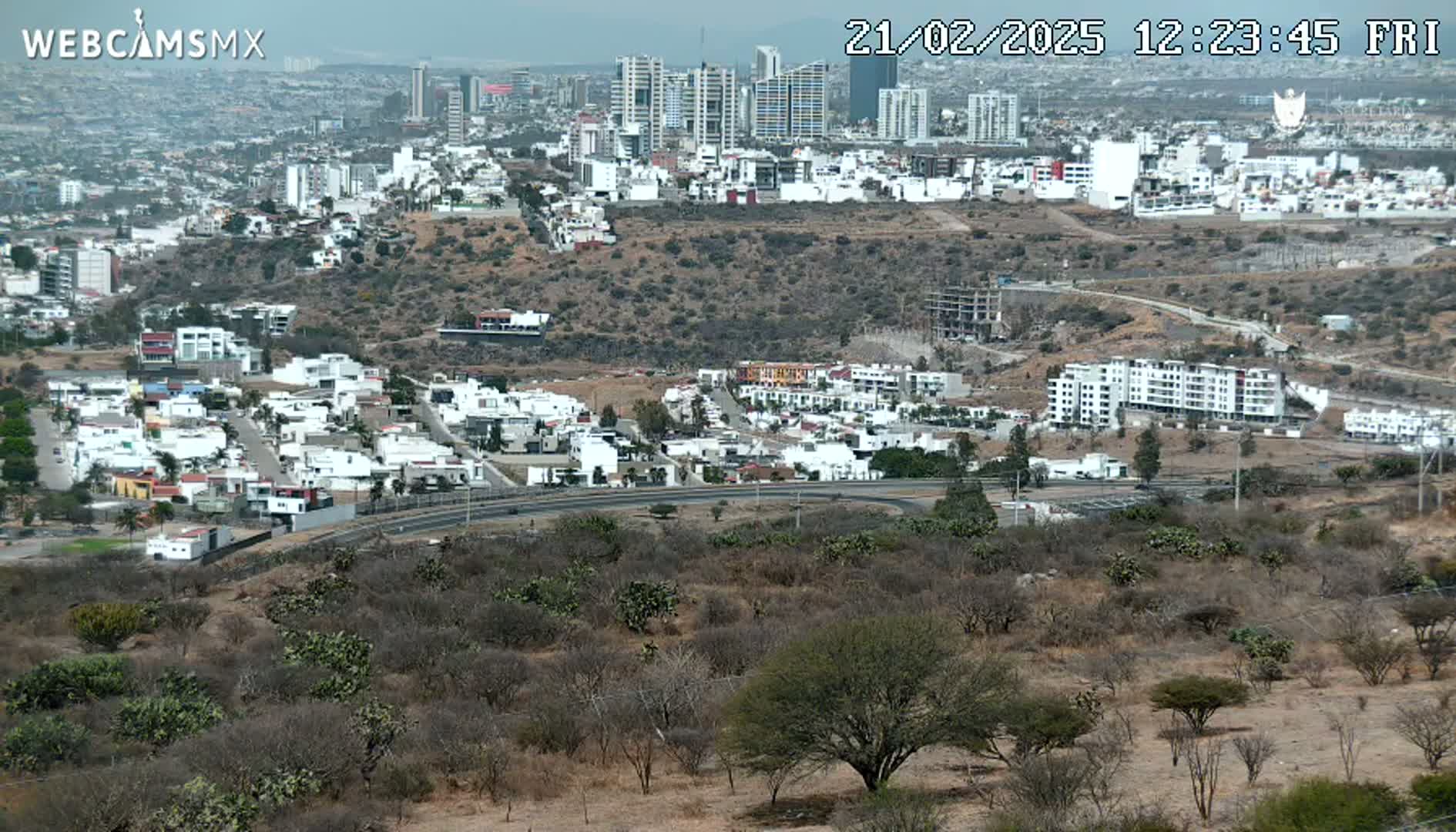 A sprawling city landscape under a hazy sky, featuring a mix of low-rise residential buildings and taller apartment complexes, with dry, scrubby vegetation in the foreground.