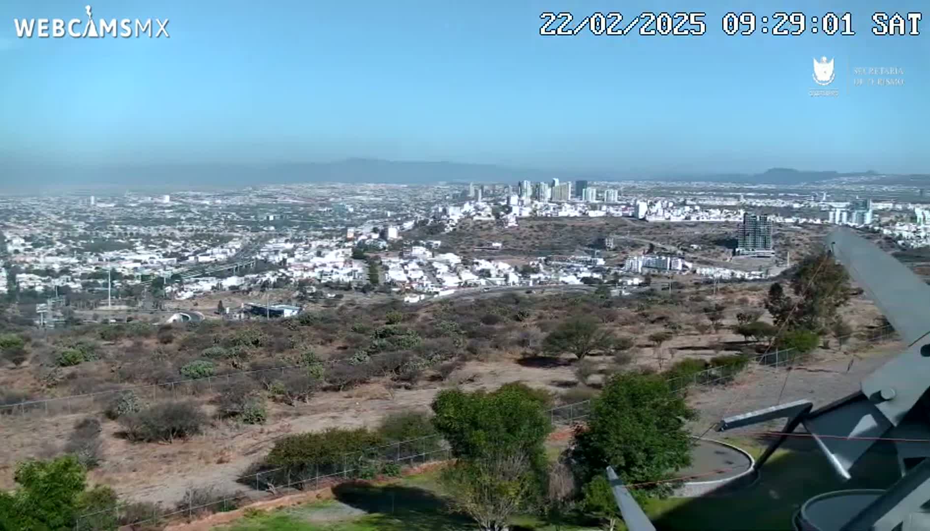 A high-angle view on a clear, sunny day shows a sprawling city with a coastal view in the distance, partially obscured by a hillside with sparse vegetation and a metal structure in the foreground.