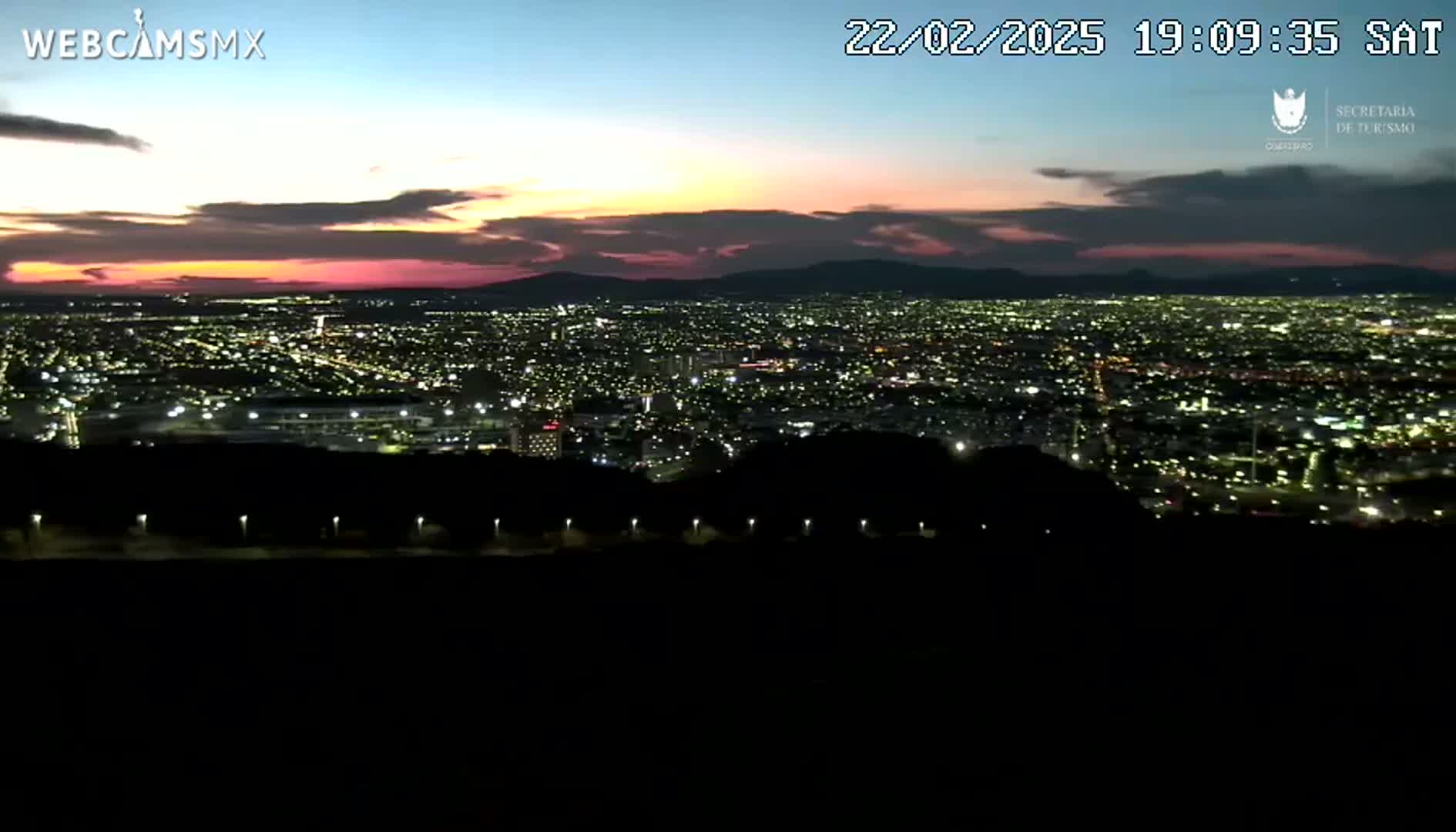 A nighttime view of a sprawling city illuminated by lights, set against a backdrop of mountains under a twilight sky with hints of pink and purple hues.