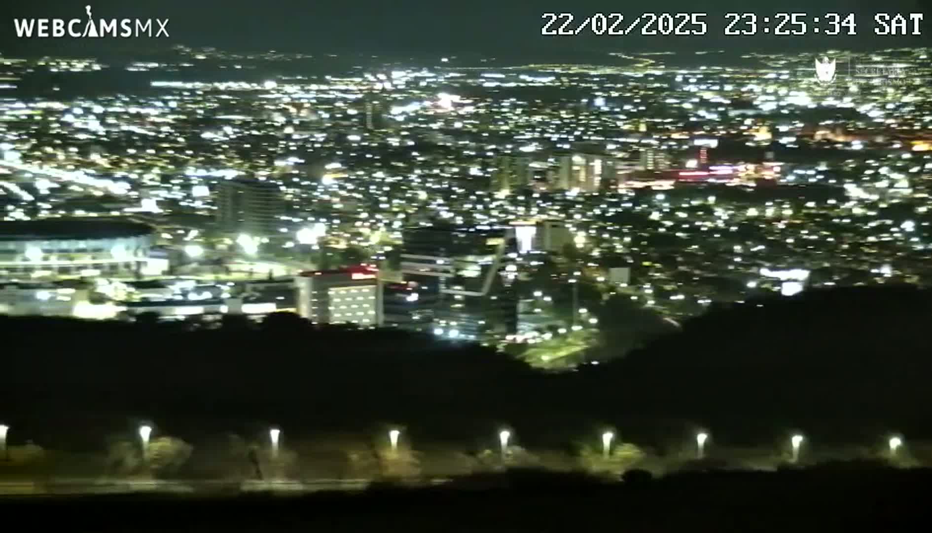 A nighttime, high-angle view shows a sprawling city illuminated by countless lights, partially obscured by a dark, silhouetted foreground.