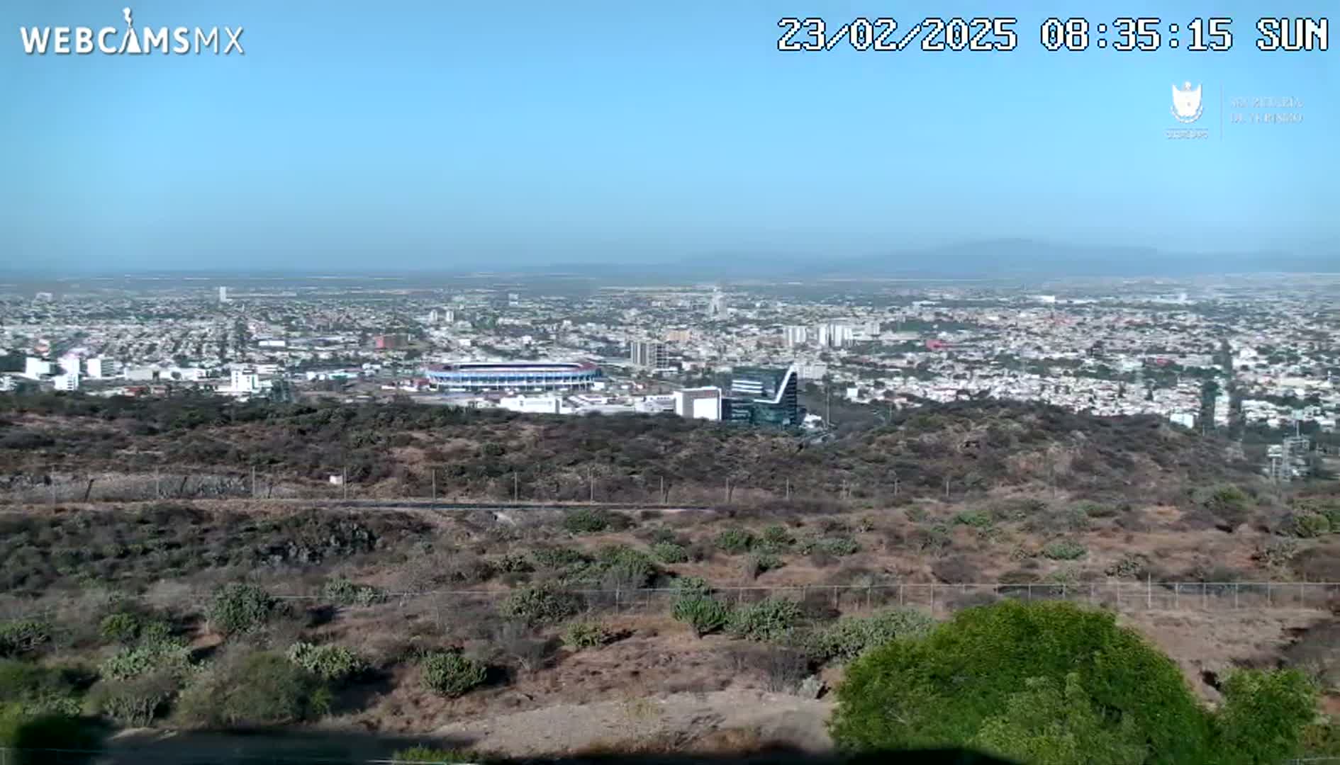 A sprawling city with a stadium is visible from a hilltop vantage point on a sunny day.