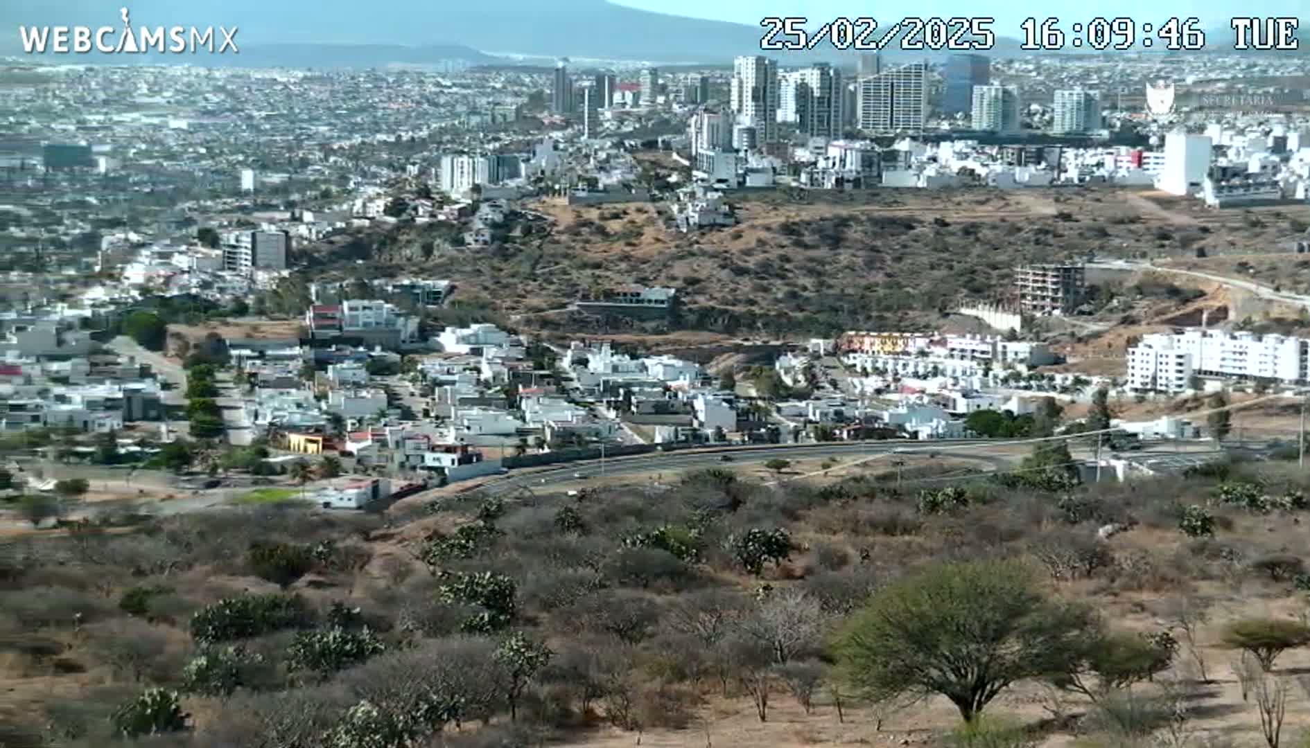 A sprawling city scene is viewed from a high vantage point on a clear, sunny day, showing residential areas, high-rise buildings, and a highway winding through a dry, scrub-covered landscape.