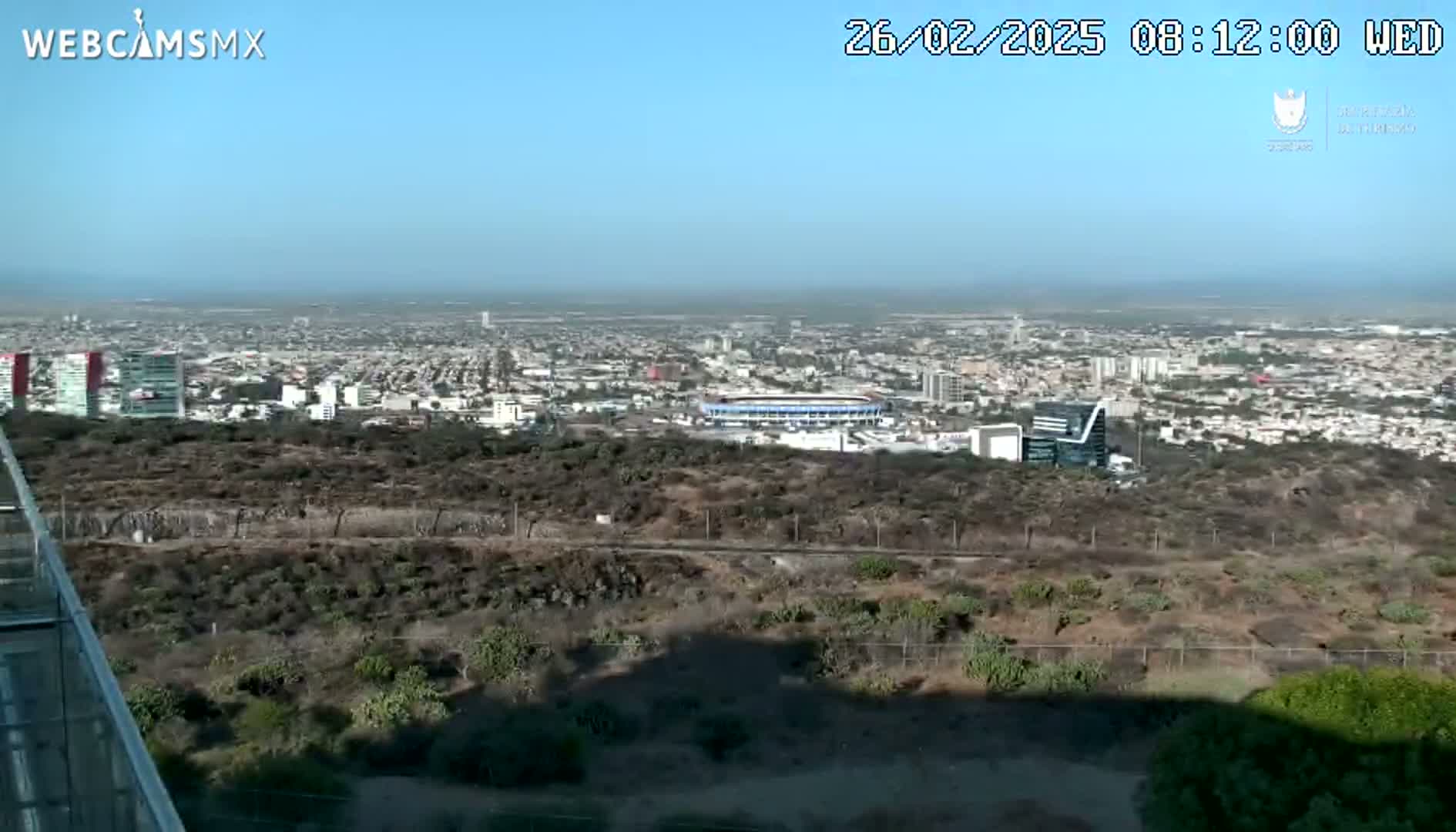 A sprawling city is visible from a high vantage point on a clear, sunny day, with scrubby vegetation in the foreground and a stadium nestled within the urban landscape.