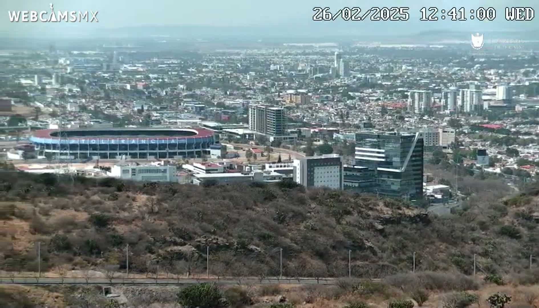 A long shot of a city on a hazy day, featuring a stadium and several buildings, viewed from a hill overlooking the urban landscape.