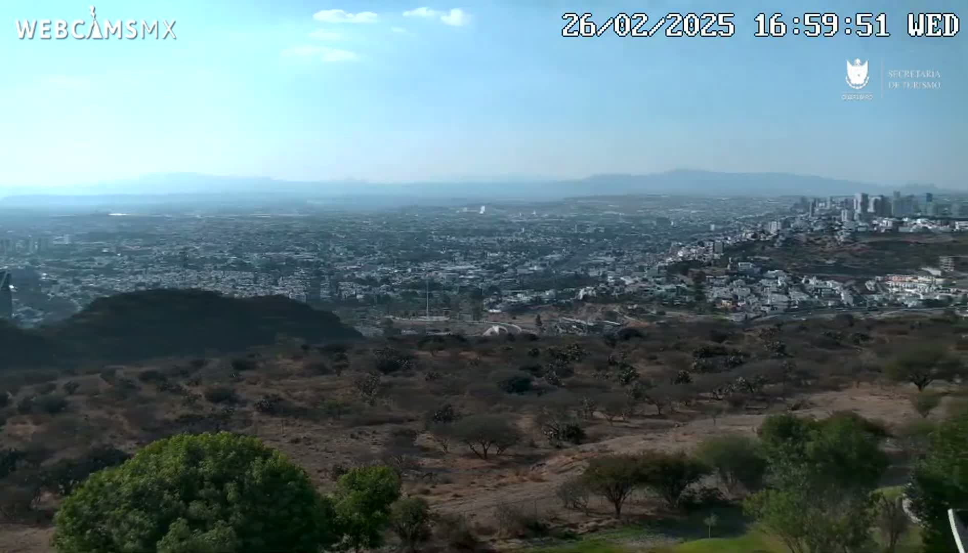 A sprawling city is visible in the distance under a mostly clear sky, viewed from a high vantage point overlooking dry, scrubby hills and sparse vegetation.