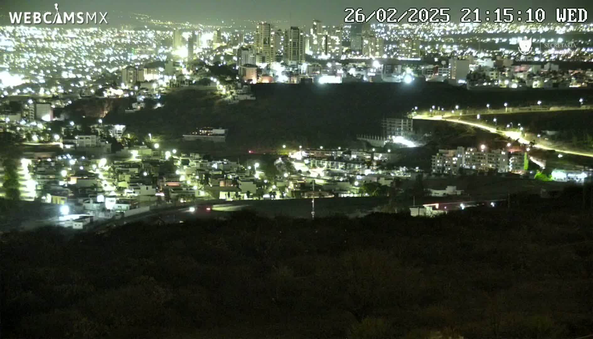 A nighttime view from a hilltop shows a city sprawling across a valley, illuminated by numerous lights.