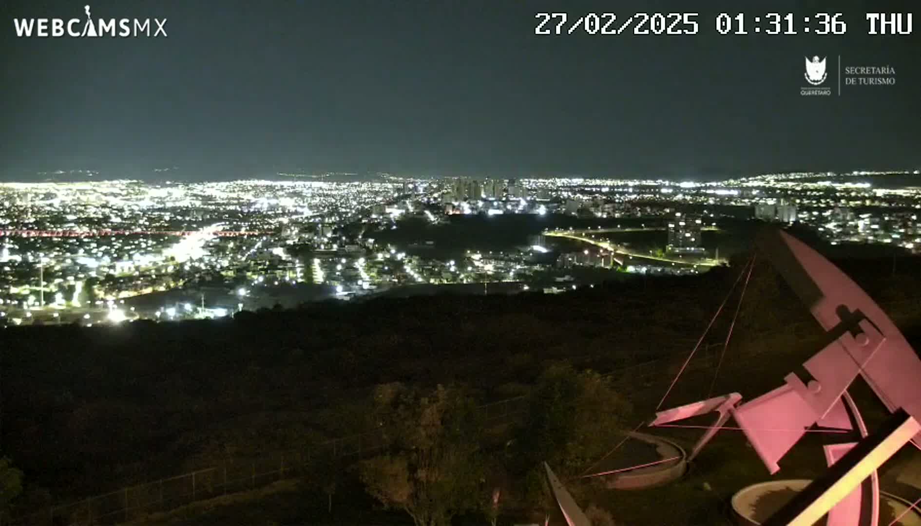 A nighttime, long shot of a city brightly lit below a hilltop, with a large metal sculpture partially visible in the foreground.